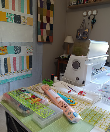 Cluttered sewing table and machine wiht unfinished quilts on design wall in background