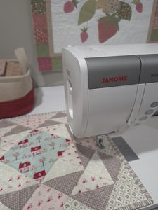 Sewing machine needle housing on table with quilted block on bed, quilted wall hanging in the background and a basket of fabrics